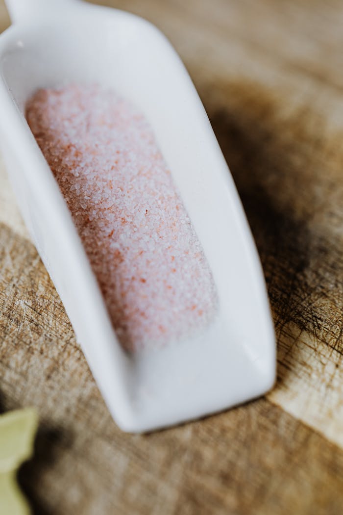 A close-up view of Himalayan pink salt in a white ceramic scooper on a wooden surface.